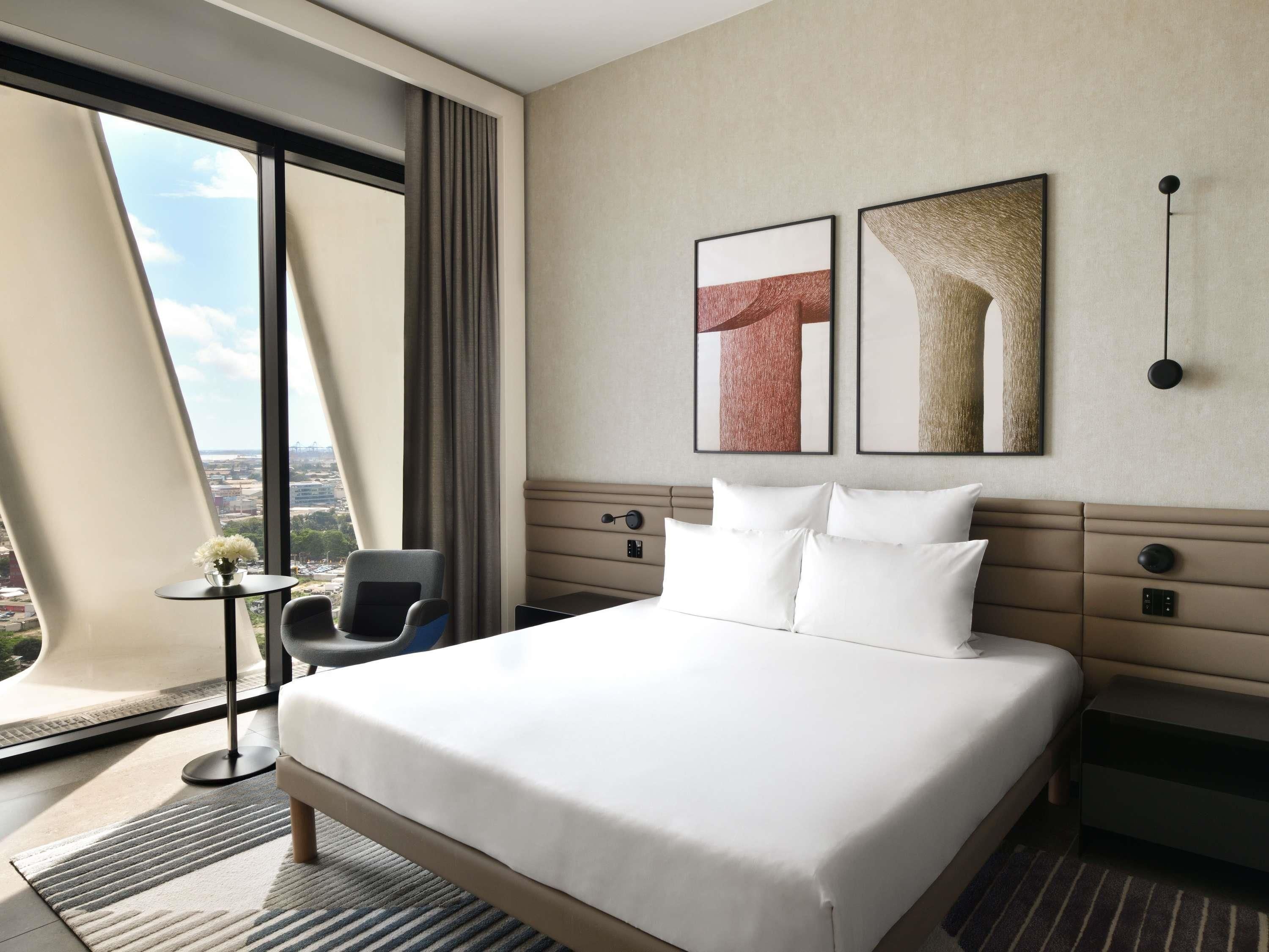 Una camera d'albergo con un letto grande, una poltrona, un tavolino e vista sulla città attraverso ampie finestre.