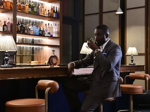 A man seated at a bar with a bottle of liquor, a lamp, and a bar counter. - Novotel Abidjan Marcory (Abidjan)