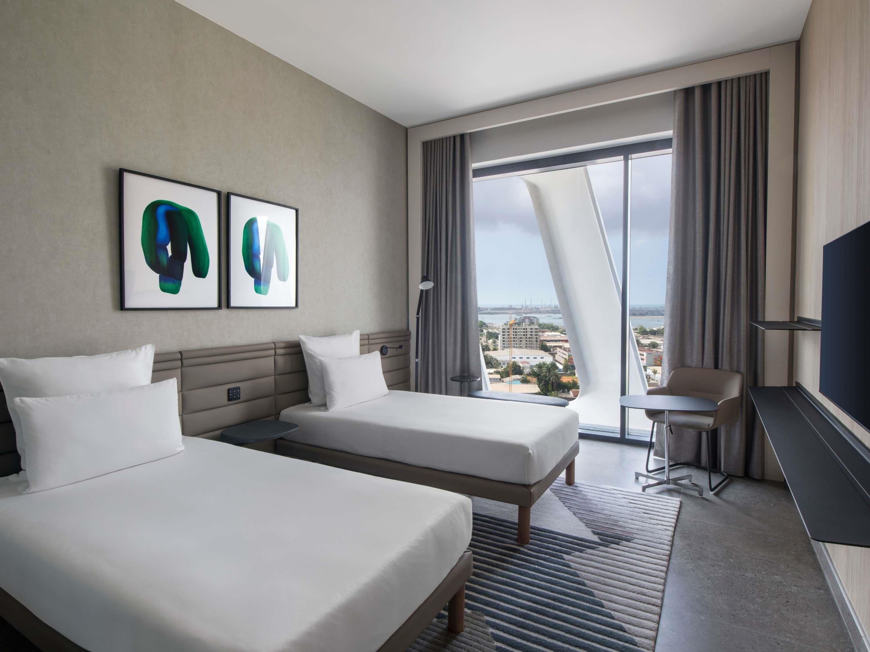 A hotel room with two beds, a desk, a chair, a TV, and a large window with a city view.