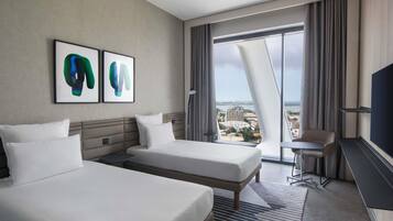 A hotel room with two beds, a desk, a chair, a TV, and a large window with a city view.