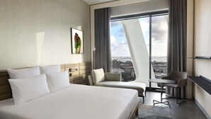 Una habitación de hotel moderna con una cama grande, dos sillones, una mesita pequeña y vistas de la ciudad a través de ventanales del suelo al techo.