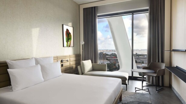 A modern hotel room with a large bed, two armchairs, a small table, and a city view through floor-to-ceiling windows. - Novotel Abidjan Marcory (Abidjan)