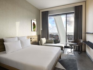 A modern hotel room with a large bed, two armchairs, a small table, and a city view through floor-to-ceiling windows. - Novotel Abidjan Marcory (Abidjan)