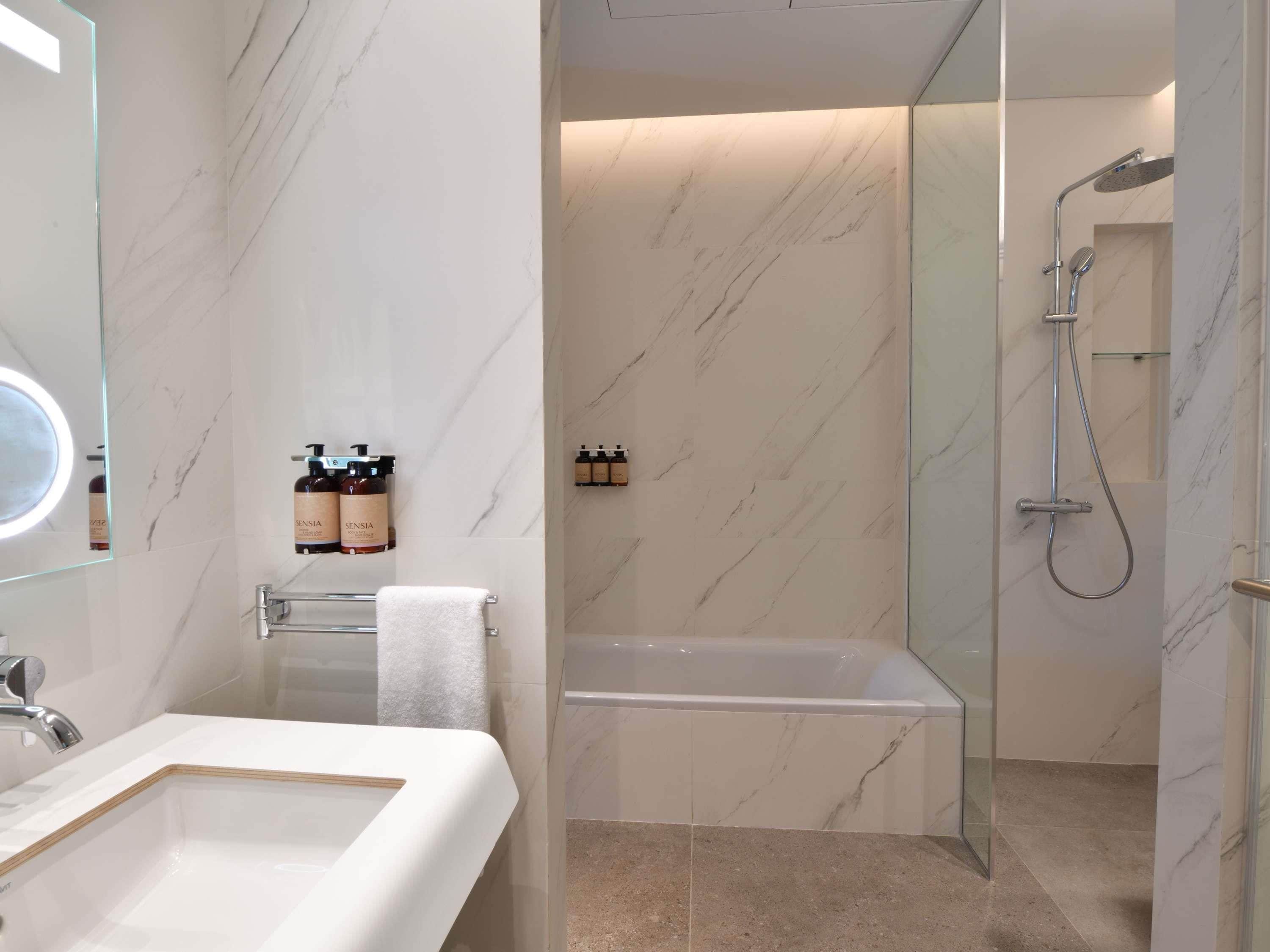 Un baño moderno con pared de mármol, ducha con cerramiento de vidrio, bañera blanca y tocador con espejo y lavabo.
