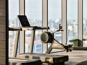 A treadmill with a laptop on a stand, a stationary bike, and a view of a cityscape through large windows. - Novotel Abidjan Marcory (Abidjan)