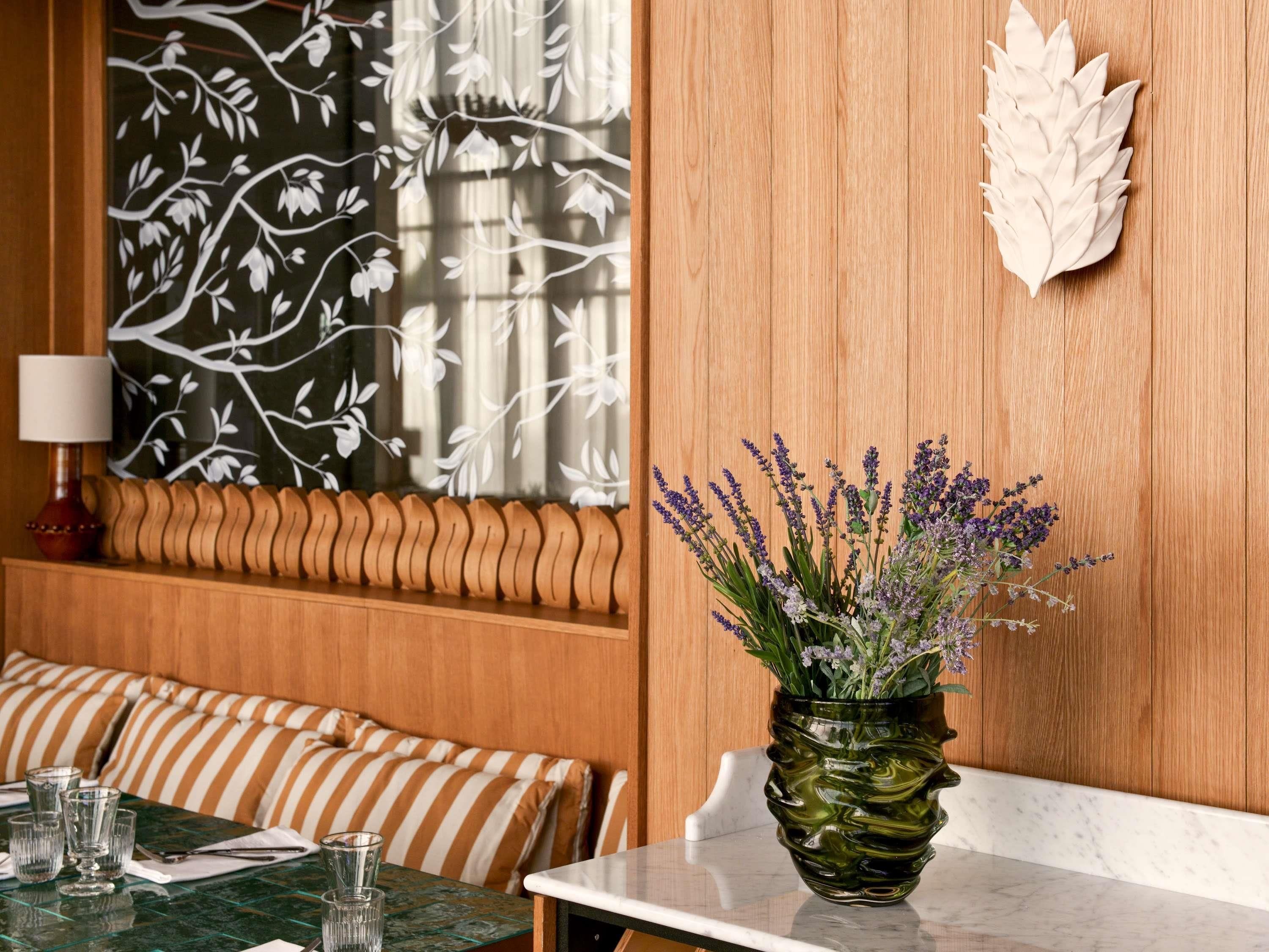 Een eetruimte met een tafel voor vier personen, een decoratieve vaas met lavendel, een houten wand met een bladmotief en een gestreept kussen.