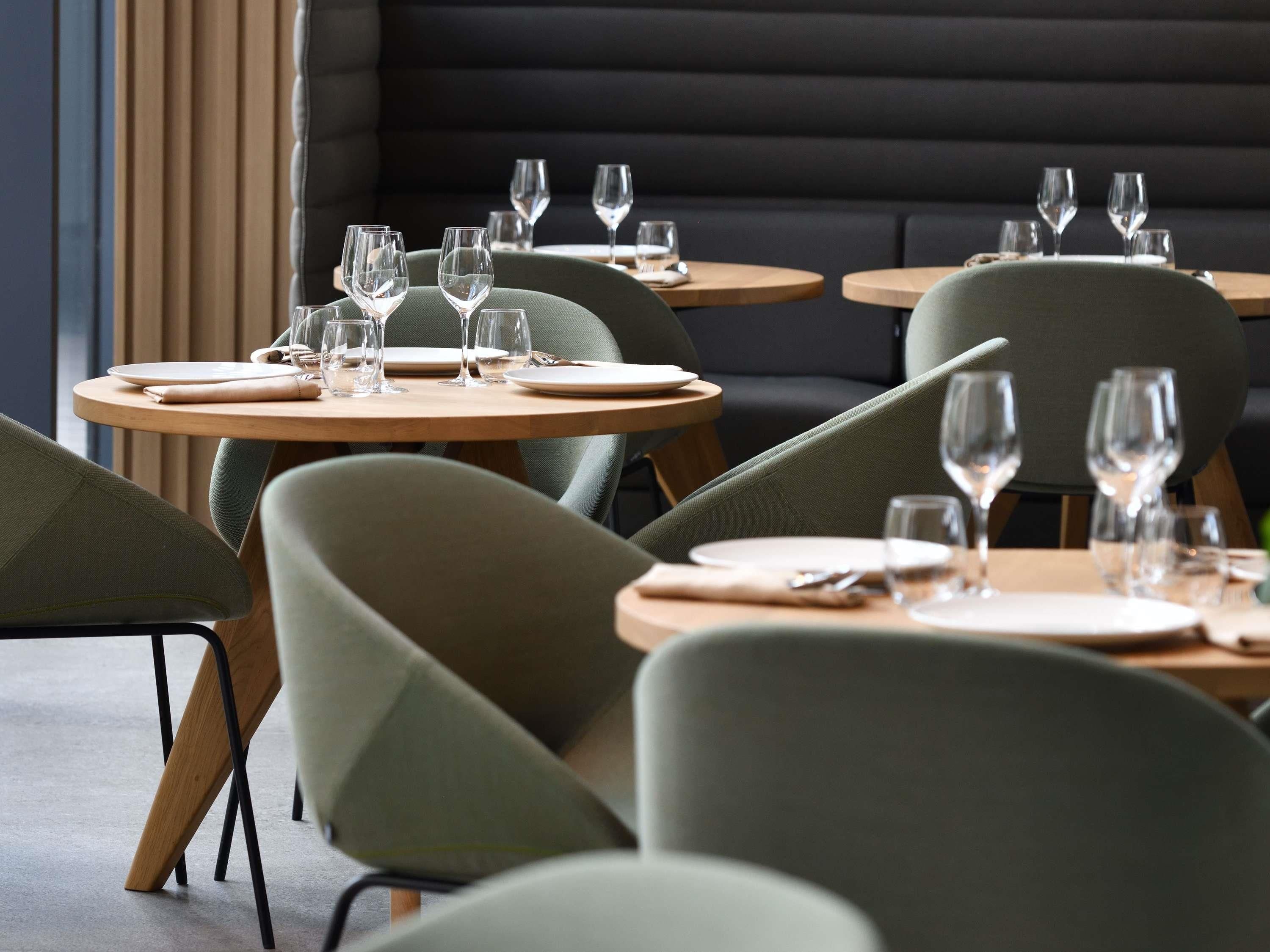 A restaurant interior with round tables, green upholstered chairs, and a dark cushioned bench in the background.