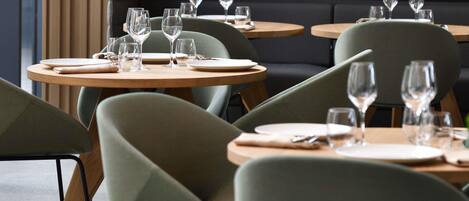 A restaurant interior with round tables, green upholstered chairs, and a dark cushioned bench in the background.