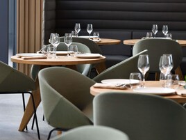 Un interior de restaurante con mesas redondas, sillas tapizadas de verde y un banco acolchado oscuro al fondo.