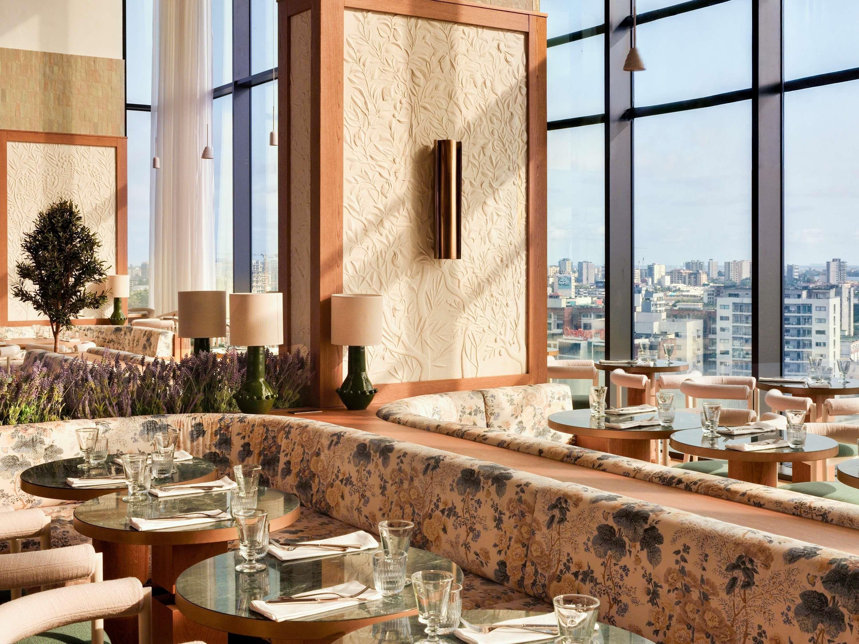 En moderne restaurant med runde borde og blomstret betræk, store vinduer der giver udsigt over byen, samt en dekorativ væg med bladmotiver.