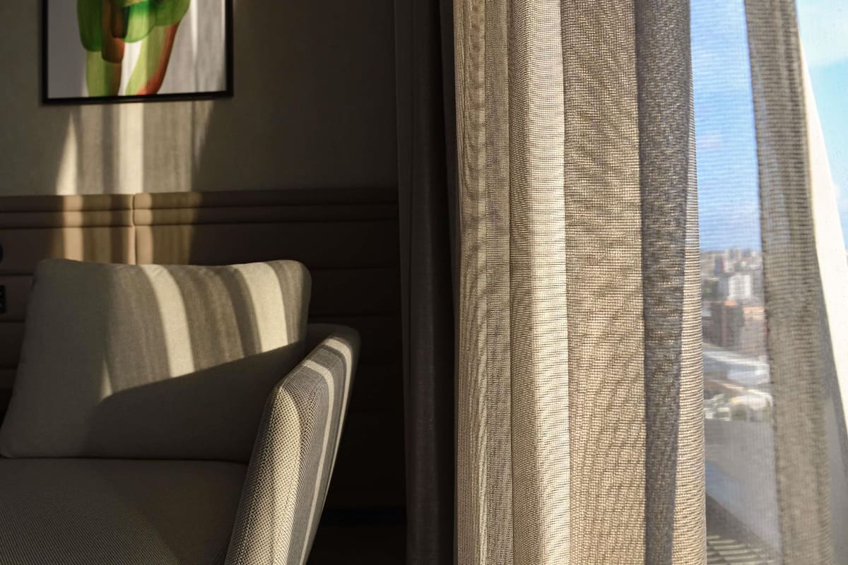 A hotel room with a sofa, a window with sheer curtains, and a cityscape view.