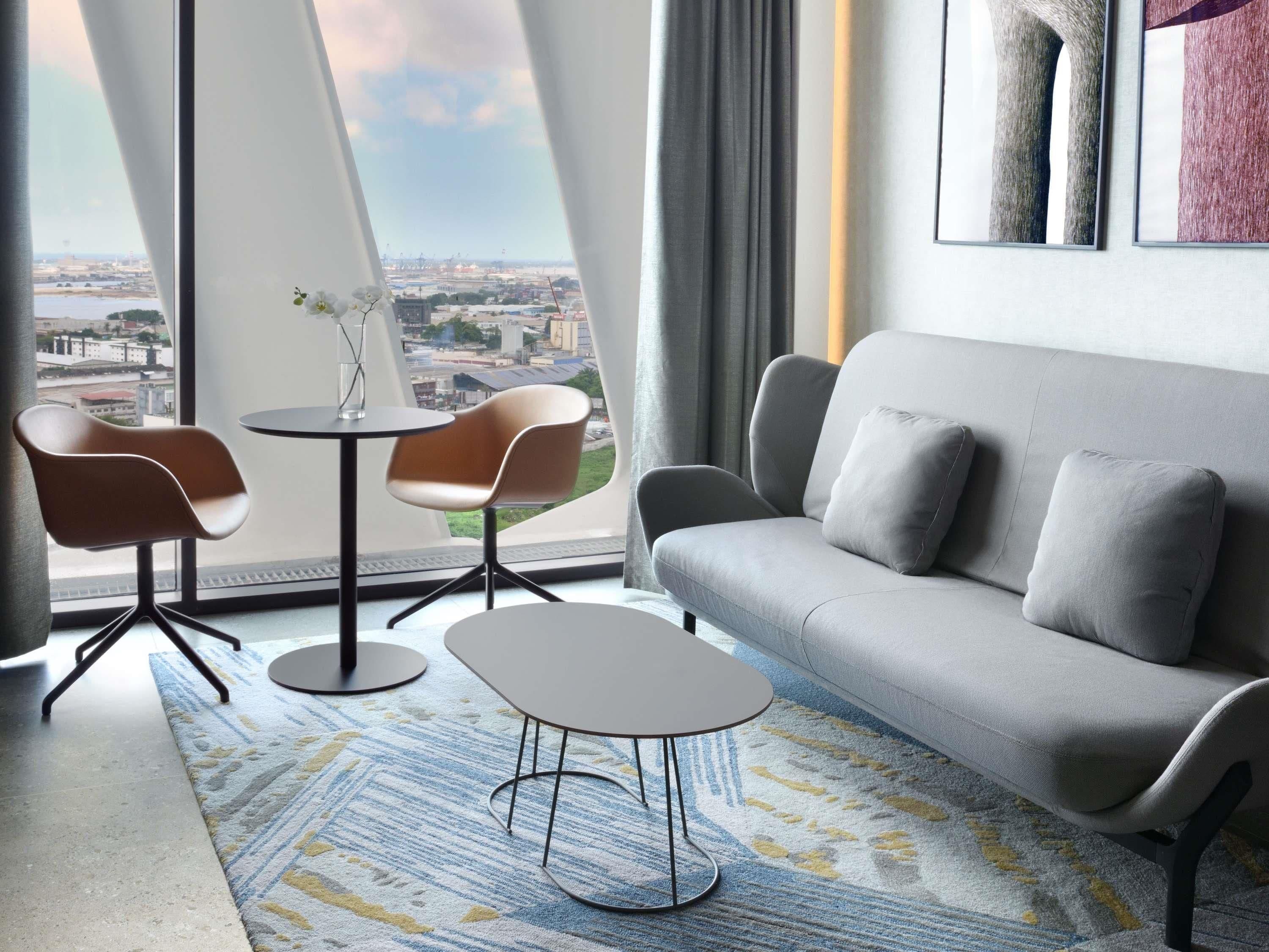 Una sala de estar moderna con un sofá gris, dos sillones, una mesita redonda y vistas a un paisaje urbano a través de grandes ventanales.