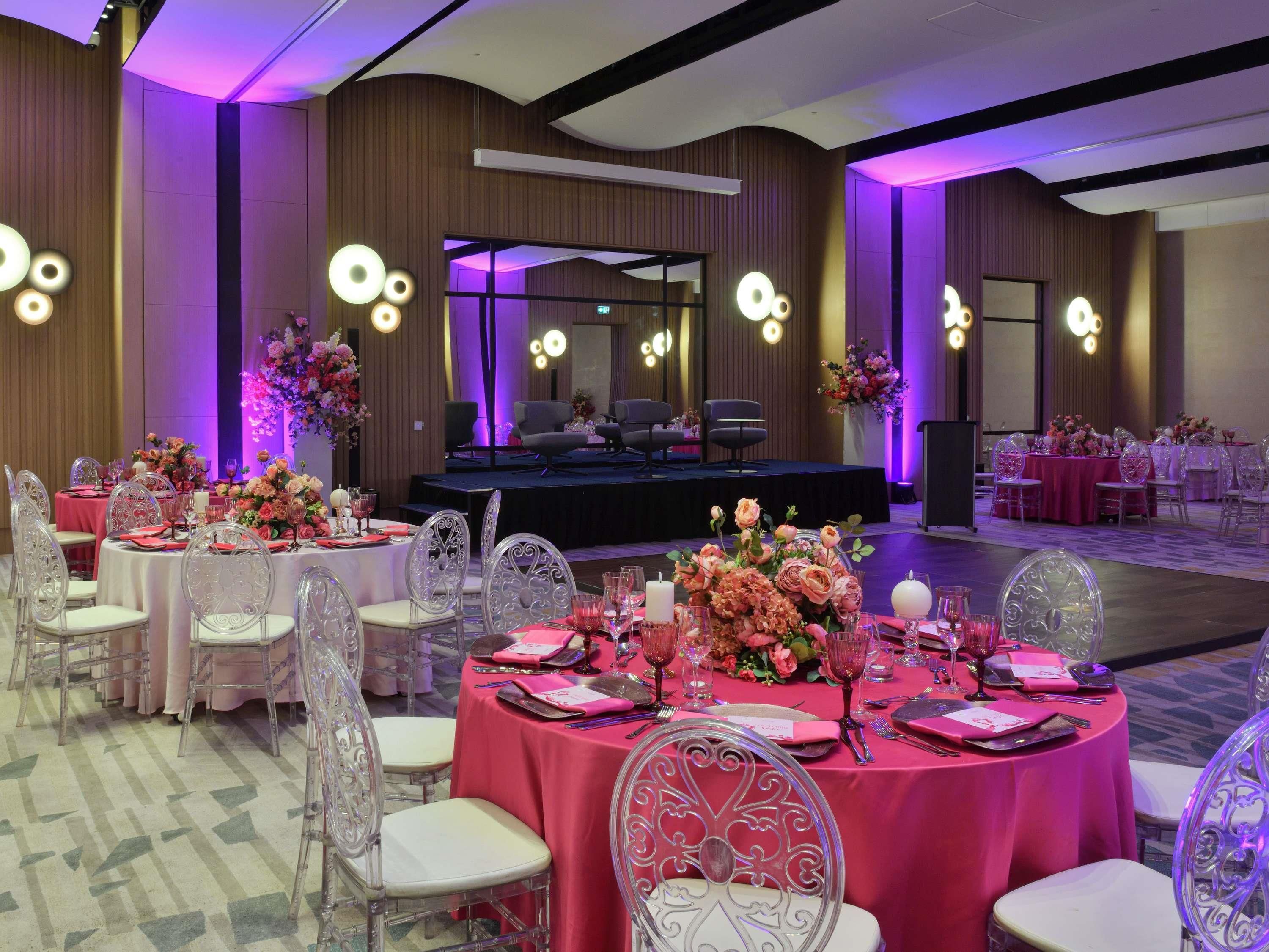 A banquet hall with round tables, clear chairs, and floral centerpieces.