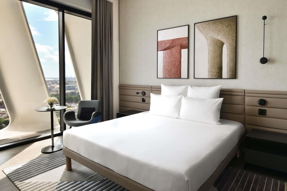 A hotel room with a large bed, a chair, a small table, and a view of the city through large windows.