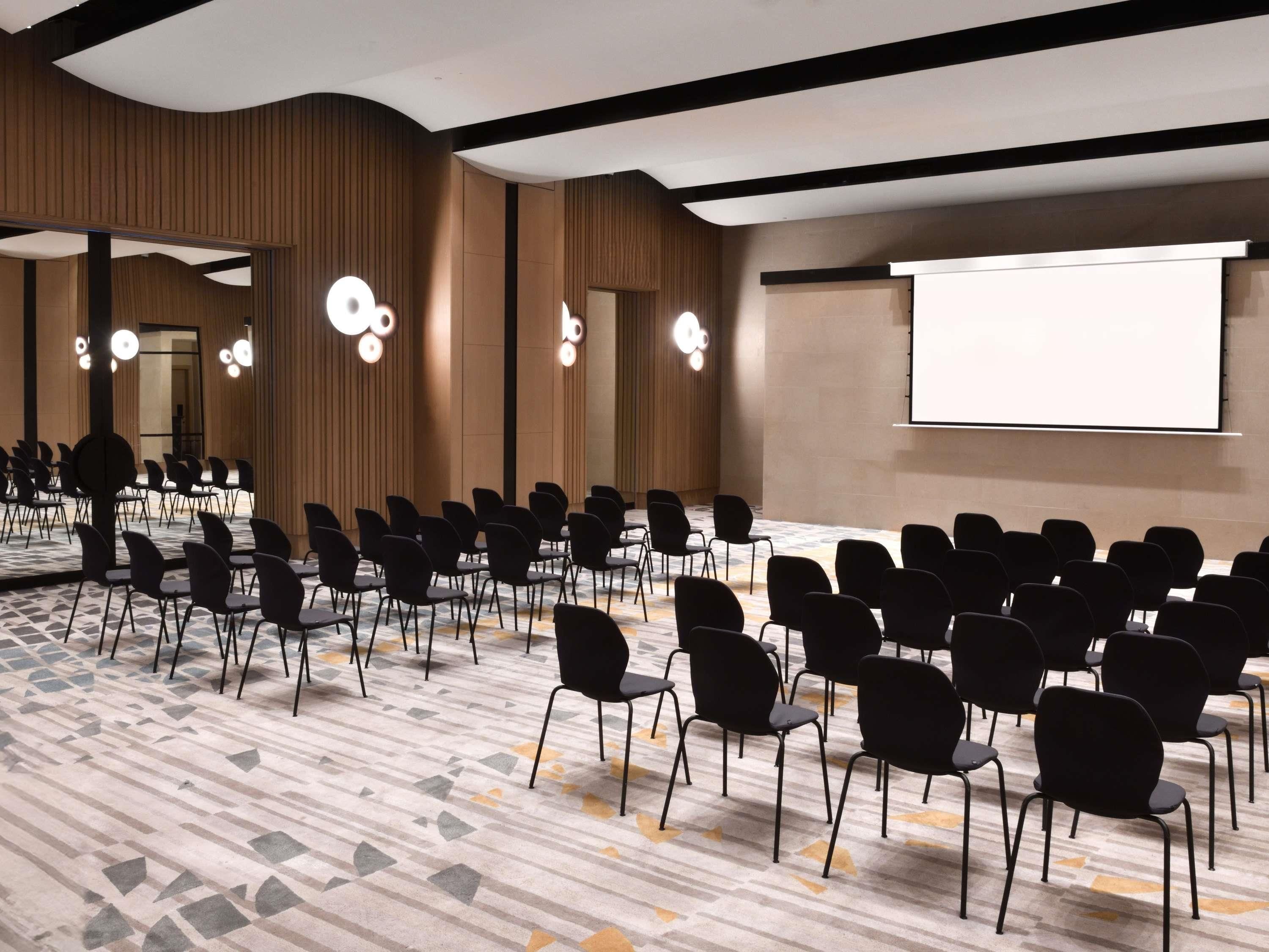 Ein Konferenzraum mit Reihen von Stühlen, einer großen Leinwand und modernen Leuchten.
