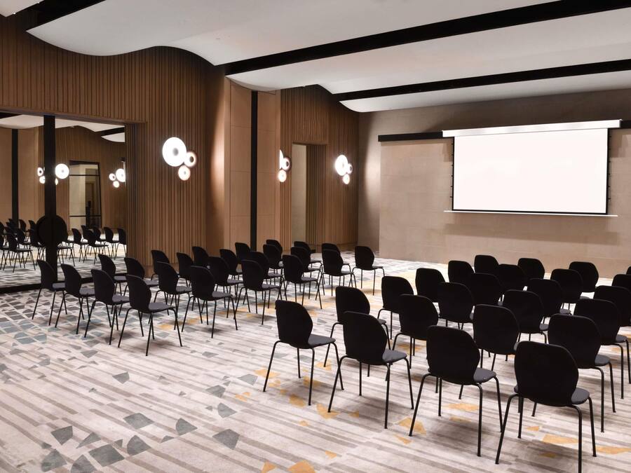 Ein Konferenzraum mit Reihen von Stühlen, einer großen Leinwand und modernen Leuchten.