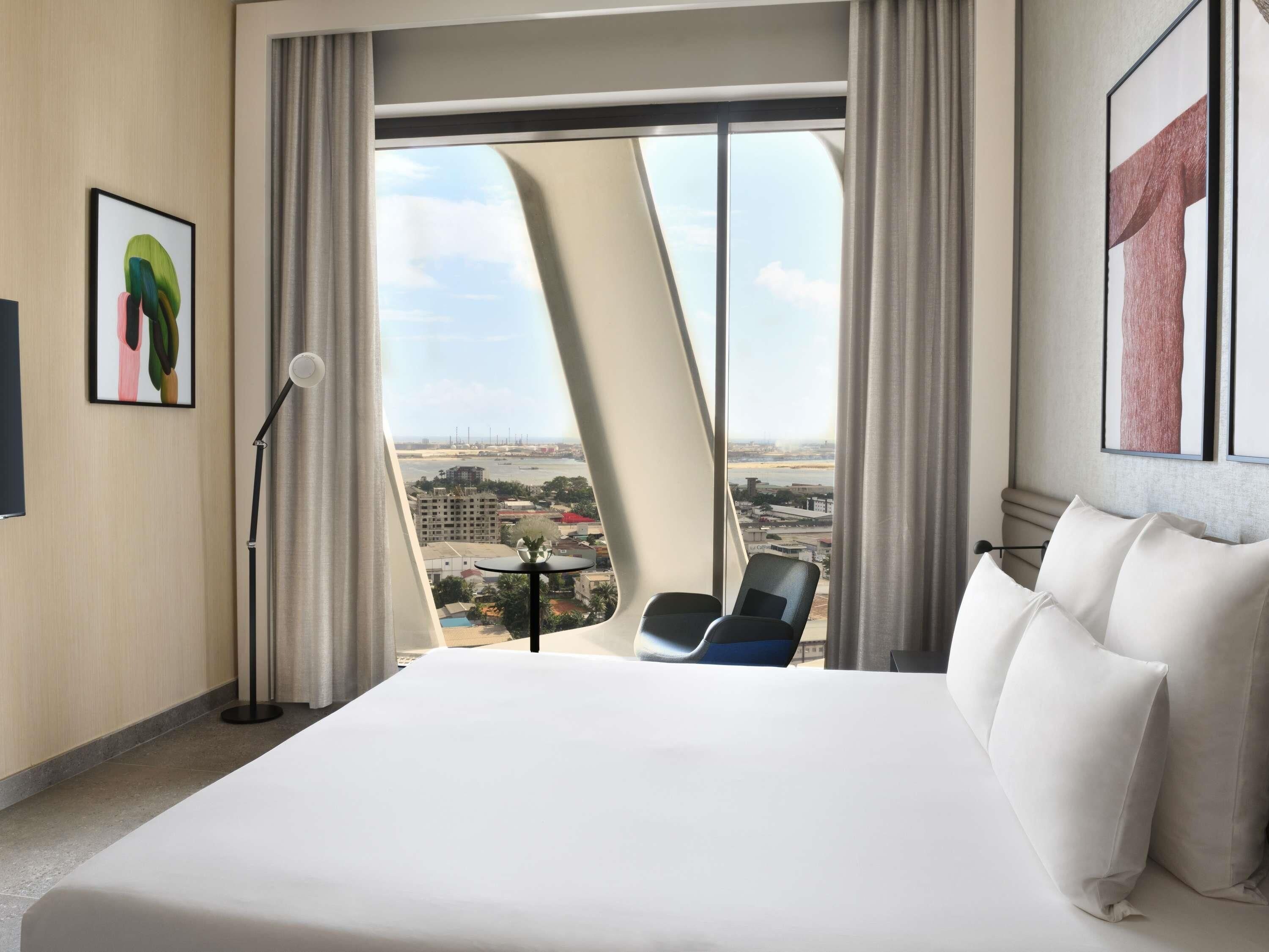 Une chambre d’hôtel avec un grand lit, une chaise, un bureau agrémenté d’un vase et une vue sur le paysage urbain.