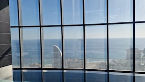 Visão através de uma grande janela com padrão de grade, com vista para uma área costeira com edifícios e céu claro.