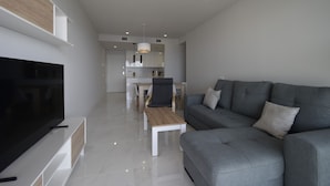 Uma sala moderna com televisão de tela plana, sofá cinza, mesa de centro de madeira e uma área de refeições ao fundo.