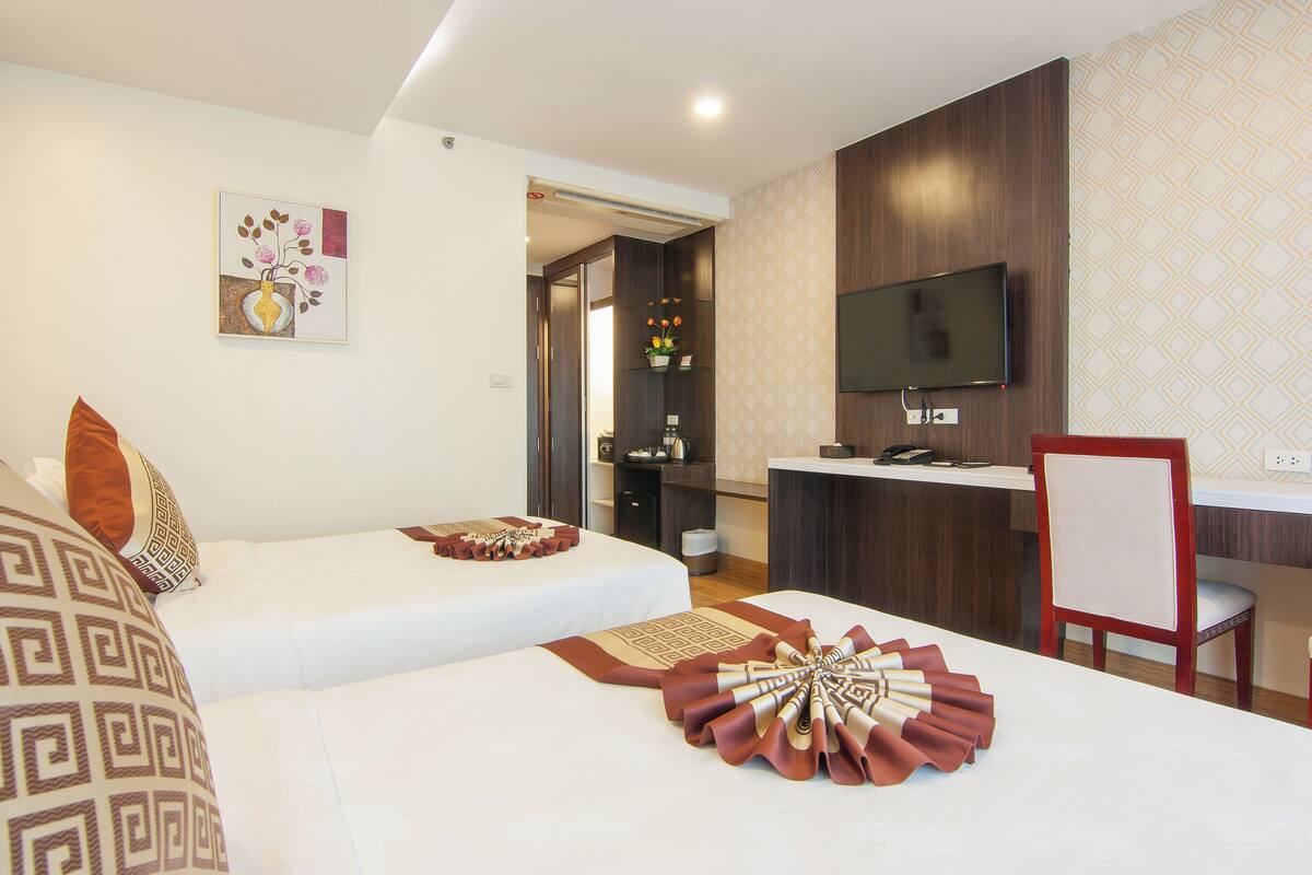 Habitación de hotel con dos camas, televisor, escritorio y baño visible a través de una puerta abierta.