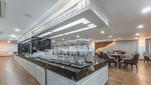 Un comedor de hotel moderno con una larga barra, varias máquinas de café y una zona de estar con mesas y sillas.