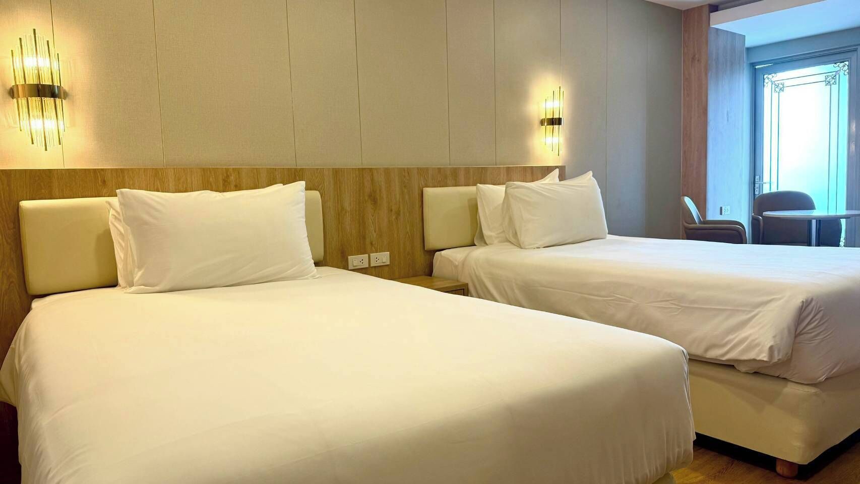 Habitación de hotel con dos camas, cabecera de madera, luces fijadas a la pared y vistas al exterior.