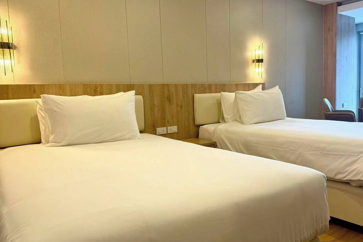 Habitación de hotel con dos camas, cabecera de madera, luces fijadas a la pared y vistas al exterior.