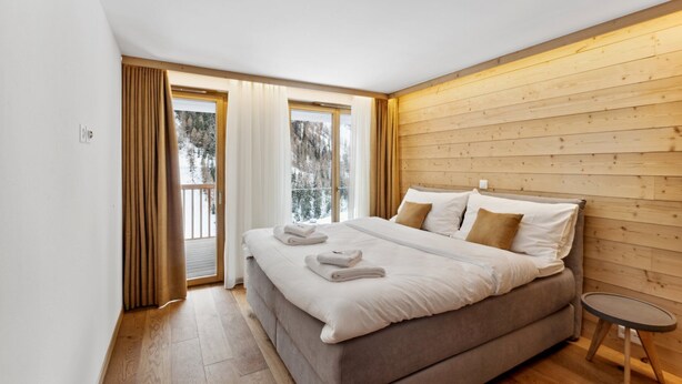 A modern bedroom with a large bed, wooden panel walls, and a view of snowy trees through the windows. - Luxury Apartment, 2 Bedrooms Rahas B Grimentz - By Mrs Miggins (Grimentz)