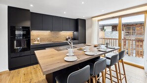A modern kitchen with a wooden table and chairs, a built-in oven, and a view of a wooden deck. - Luxury Apartment, 2 Bedrooms Rahas B Grimentz - By Mrs Miggins (Grimentz)