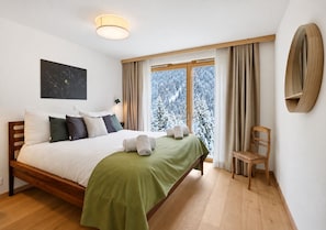 A bedroom with a wooden bed, a green bedspread, a window with curtains, a chair, and a wall-mounted shelf.
