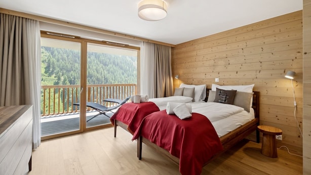 Un dormitorio con cama, una mesita de noche, una lámpara, suelo de madera, un balcón con vista a un bosque y una luz en el techo.