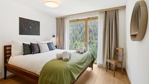 Un dormitorio con cama, mesita de noche, silla, ventana con cortinas y una lámpara fijada a la pared.