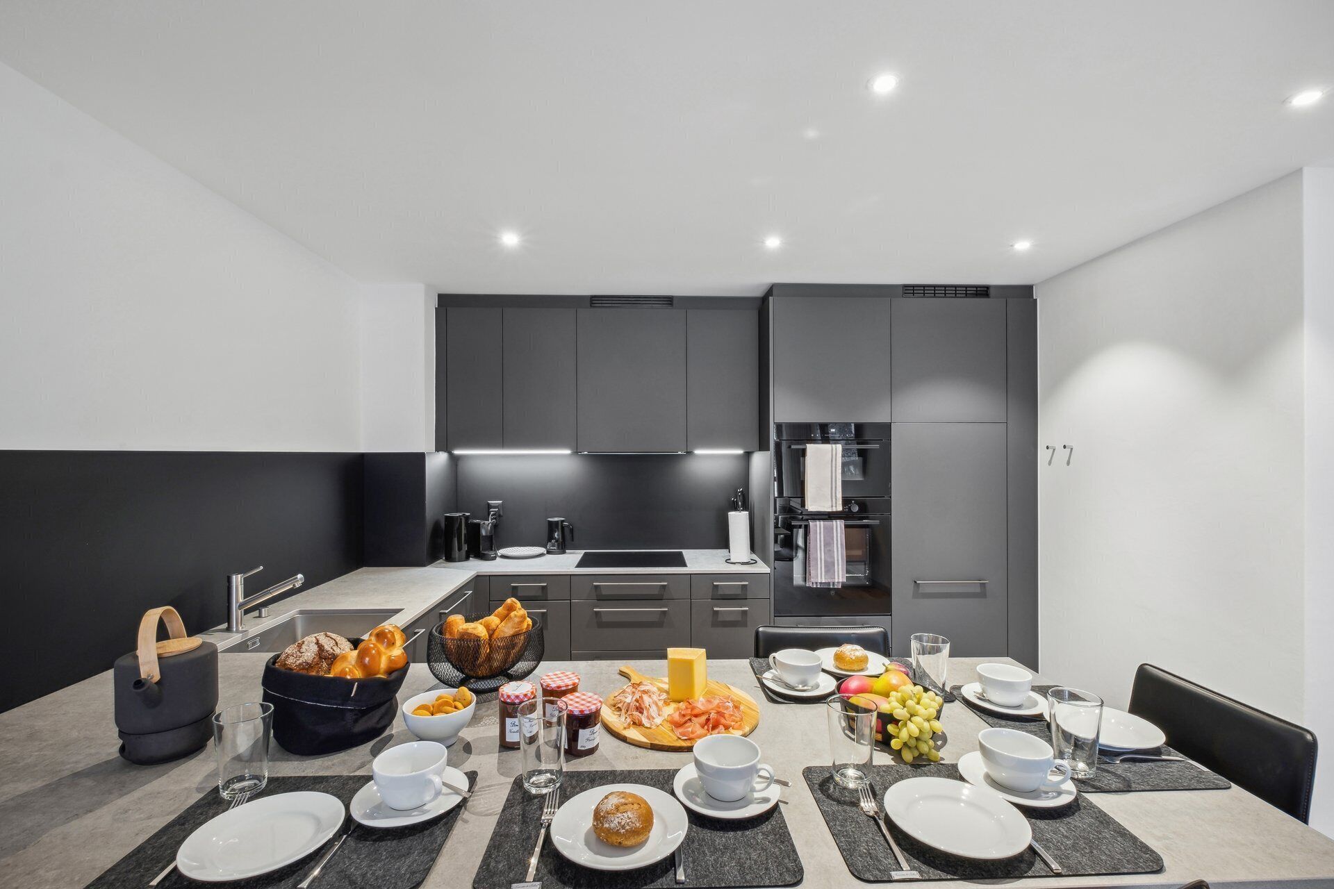 Una cucina moderna con mobili grigi, un'area pranzo apparecchiata per la colazione con diversi cibi e bevande, e un frigorifero sullo sfondo.