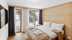 Ett sovrum med en sänggavel i trä, en stor säng med vita sängkläder, ett nattduksbord i trä och en utsikt över ett snötäckt landskap genom fönstren.