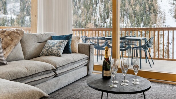 Ett mysigt vardagsrum med en soffa, kuddar, ett litet runt bord med en flaska och glas, och utsikt över en snötäckt skog genom stora fönster.
