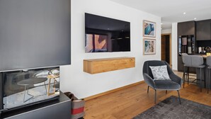 Ett modernt vardagsrum med en platt-TV, ett bord i trä, en grå fåtölj och en matplats med barstolar.