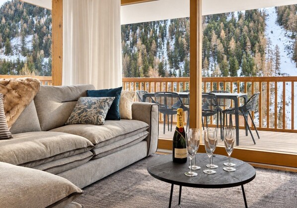 A cozy living area with a sofa, a table with a bottle and glasses, and a view of a forested mountain.