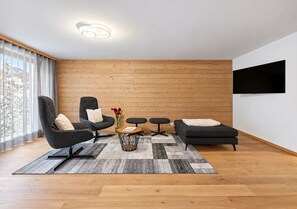 A modern living room with a wooden wall, a flat-screen TV mounted on the wall, a grey sofa, a black armchair, and a patterned rug.