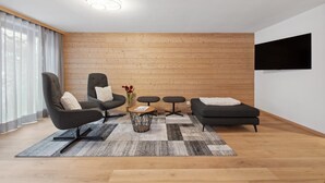 Uma sala de estar moderna com painel de madeira, um sofá preto, uma poltrona cinza, uma mesa de centro e uma TV na parede.