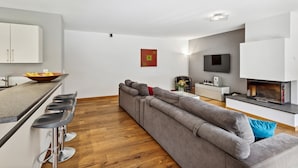 A modern living room with a grey sectional sofa, a kitchen area with white cabinets, a dining bar with stools, a flat-screen TV mounted on the wall, and a fireplace. - Beautiful apartment at The Lodge Champéry - By Mrs Miggins (Champéry)