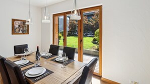 A dining area with a wooden table set for six, large windows with a view of a garden, and pendant lighting. - Beautiful apartment at The Lodge Champéry - By Mrs Miggins (Champéry)
