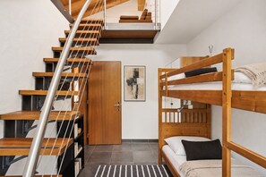 A dormitory room with bunk beds, a wooden staircase, and a door leading to another room.