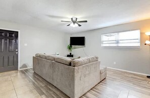 A living room with a sectional sofa, a ceiling fan, a flat-screen TV, and a window with blinds. - Charming 2BR Apartment in Central Lawton Near Fort Sill (Lawton)