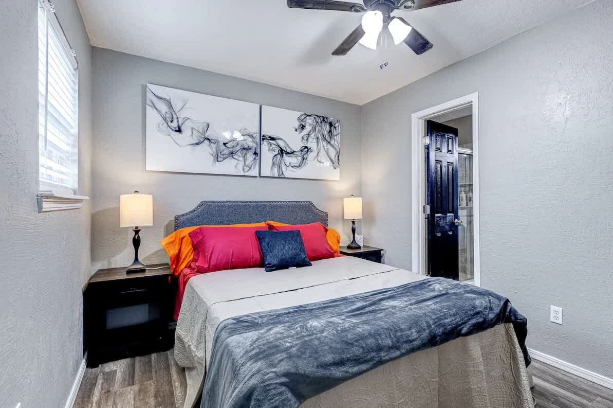 A bedroom with a bed, two bedside tables, a ceiling fan, artwork on the wall, and a door to the right.