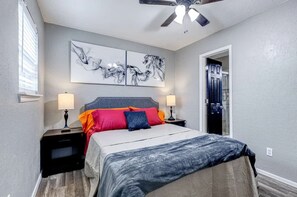 Un dormitorio con una cama, dos mesitas de noche, un ventilador de techo, una obra de arte en la pared y una puerta a la derecha.
