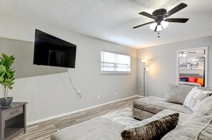 Una sala de estar con una televisión de pantalla plana montada en la pared, un ventilador de techo, una ventana con persianas, una lámpara de pie y un sofá.