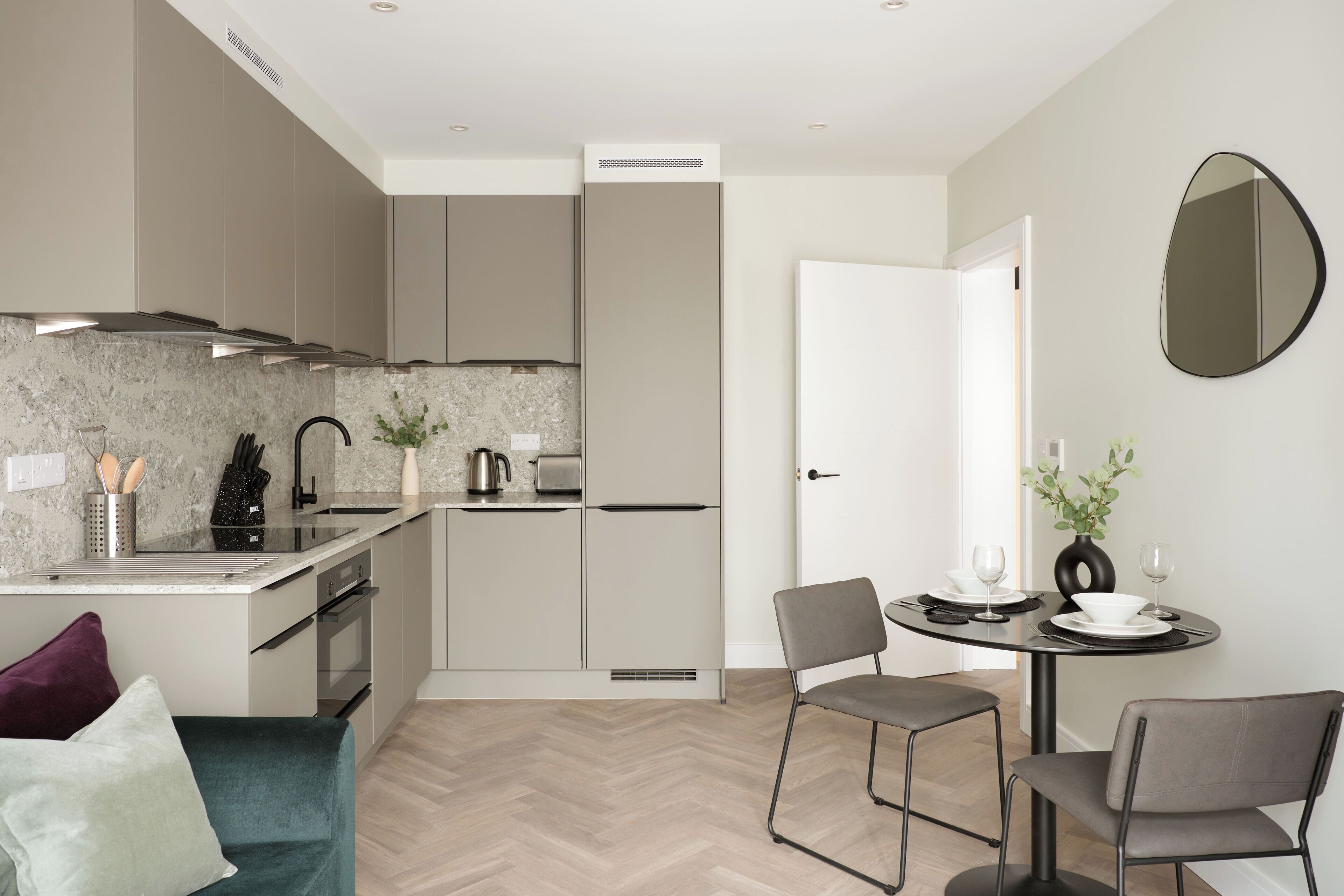 A modern kitchen with a herringbone floor, a dining area with a round table and two chairs, and a large mirror on the wall.