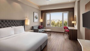A hotel room with a large bed, a sofa, a desk, and a chair. There is a window with a view of buildings and trees. - Ramada by Wyndham Ahmedabad Narendra Modi Stadium Motera (Ahmedabad)