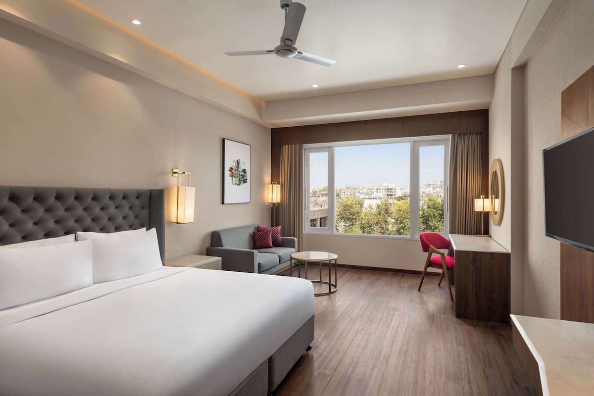 A hotel room with a large bed, a sofa, a desk, and a chair. There is a window with a view of buildings and trees.