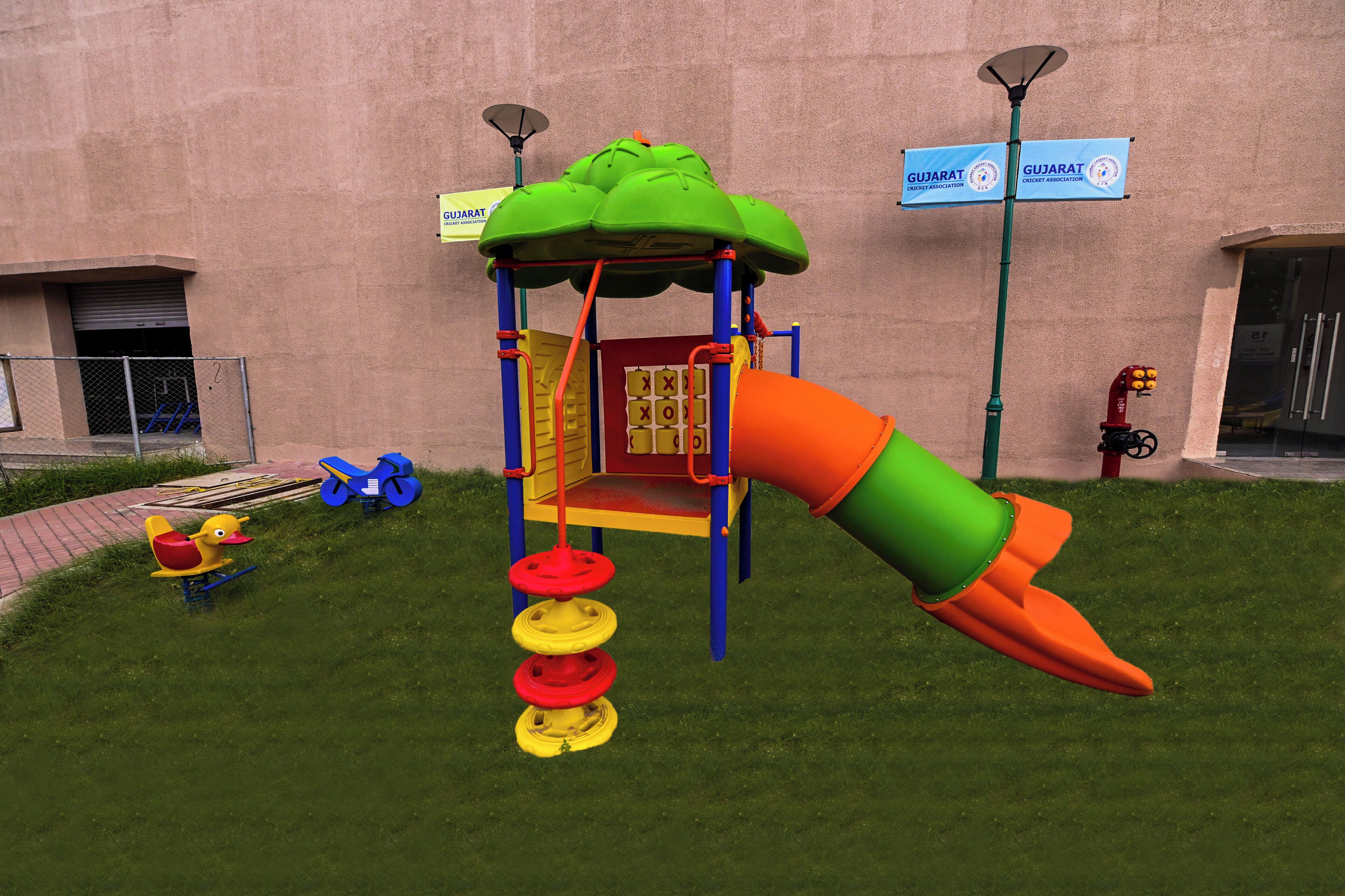 A playground with a slide and climbing elements, set against a building with signage for Gujarat Cricket Association.